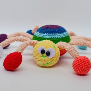 Pu&ograve; includere: Un giocattolo ragno all'uncinetto colorato con un viso giallo, occhi neri e un sorriso verde. Il ragno ha otto gambe con palline di diversi colori alle estremit&agrave;, tra cui rosso, bianco, viola e giallo.