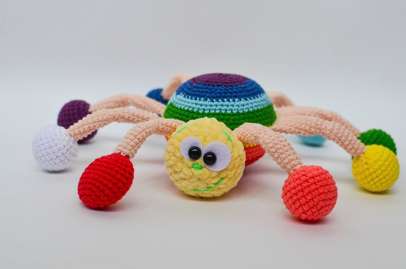 Pu&ograve; includere: Un giocattolo ragno all'uncinetto colorato con un viso giallo, occhi neri e un sorriso verde. Il ragno ha otto gambe con palline di diversi colori alle estremit&agrave;, tra cui rosso, bianco, viola e giallo.