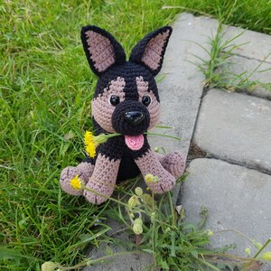 May include: A crocheted stuffed dog toy with black and brown fur, holding a yellow flower in its mouth. The dog is sitting on a bed of green grass.