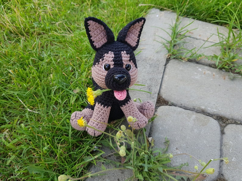 May include: A crocheted stuffed dog toy with black and brown fur, holding a yellow flower in its mouth. The dog is sitting on a bed of green grass.