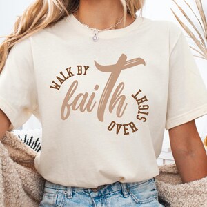 May include: A white t-shirt with a brown graphic that says "Walk by faith over sight" with a cross design.
