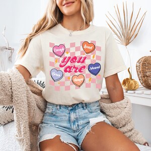 Puede incluir: Una camiseta blanca con un patr&oacute;n de cuadros rosa y blanco y corazones coloridos con palabras como "Brave", "Enough", "Sweet", "Assured", "Unique" y "You are" impresas en ella.