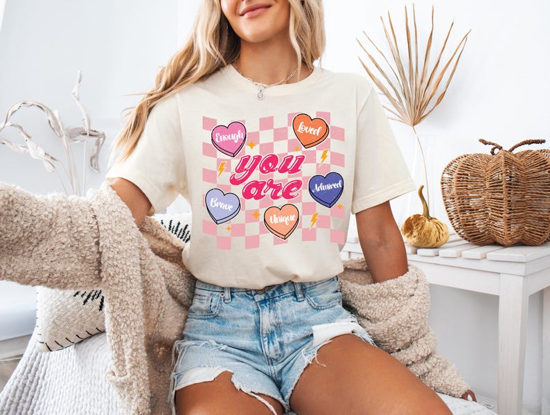 Puede incluir: Una camiseta blanca con un patr&oacute;n de cuadros rosa y blanco y corazones coloridos con palabras como "Brave", "Enough", "Sweet", "Assured", "Unique" y "You are" impresas en ella.