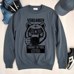 May include: A grey sweatshirt featuring a black bear graphic wearing sunglasses and holding a beer mug. The text on the sweatshirt reads "VERLOREN" and "TRINKT ZU VIEL". Additional text is present below the bear graphic.