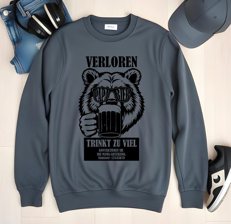 May include: A grey sweatshirt featuring a black bear graphic wearing sunglasses and holding a beer mug. The text on the sweatshirt reads "VERLOREN" and "TRINKT ZU VIEL". Additional text is present below the bear graphic.