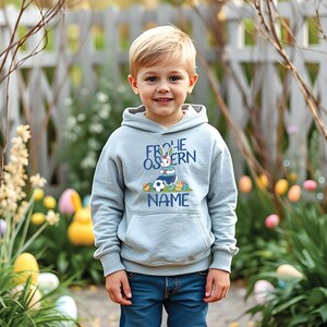 May include: A light blue hooded sweatshirt with a graphic of a bunny rabbit playing soccer and the text "Frohe Ostern" and "NAME".