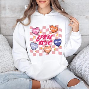 Puede incluir: Sudadera blanca con capucha con un patr&oacute;n de cuadros rosa y blanco y corazones con palabras como "Loved", "Brave", "Unique", "Enough" y "Adored". El texto "You are" est&aacute; escrito en letras rosas.