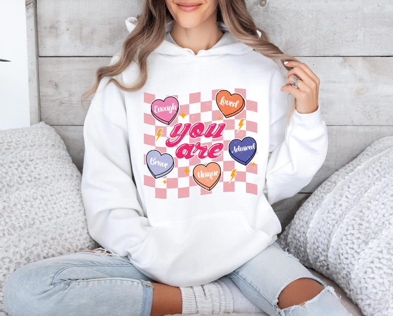 Puede incluir: Sudadera blanca con capucha con un patr&oacute;n de cuadros rosa y blanco y corazones con palabras como "Loved", "Brave", "Unique", "Enough" y "Adored". El texto "You are" est&aacute; escrito en letras rosas.