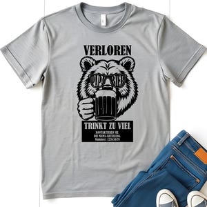 May include: A light grey t-shirt with a black graphic of a bear wearing sunglasses and holding a beer mug. The text on the shirt reads "VERLOREN", "PAPA BIER", and "TRINKT ZU VIEL". Below the bear is a black rectangle with additional text and a phone number.
