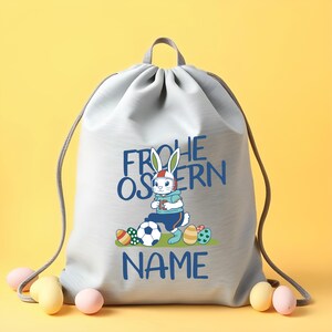 May include: A gray drawstring bag with a white bunny wearing a helmet and holding a soccer ball. The bag has the text "Frohe Ostern" and "NAME" printed on it.