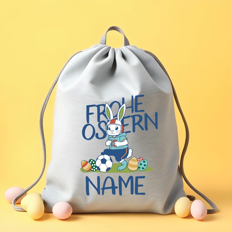 May include: A gray drawstring bag with a white bunny wearing a helmet and holding a soccer ball. The bag has the text "Frohe Ostern" and "NAME" printed on it.