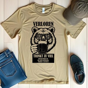 May include: A tan t-shirt featuring a black graphic of a bear wearing sunglasses and holding a beer mug. The text above the bear reads "VERLOREN" and below the bear, "TRINKT ZU VIEL". Additional text is in a black rectangle.