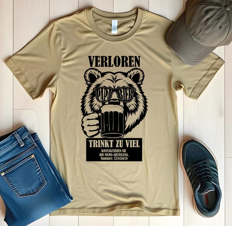May include: A tan t-shirt featuring a black graphic of a bear wearing sunglasses and holding a beer mug. The text above the bear reads "VERLOREN" and below the bear, "TRINKT ZU VIEL". Additional text is in a black rectangle.