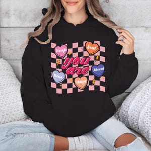 Puede incluir: Sudadera negra con capucha con un patr&oacute;n de cuadros rosa y blanco y corazones con palabras como "Amado", "Valiente", "Suficiente", "Adorado" y "&Uacute;nico". El texto "T&uacute; eres" est&aacute; en letras rosas en la sudadera.