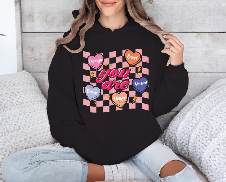 Puede incluir: Sudadera negra con capucha con un patr&oacute;n de cuadros rosa y blanco y corazones con palabras como "Amado", "Valiente", "Suficiente", "Adorado" y "&Uacute;nico". El texto "T&uacute; eres" est&aacute; en letras rosas en la sudadera.