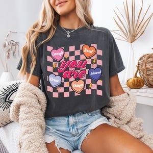 Puede incluir: Una camiseta gris oscuro recortada con un fondo a cuadros rosa y blanco. Las palabras "you are" est&aacute;n en rosa y rodeadas de cinco corazones con diferentes colores y palabras: "enough", "loved", "brave", "unique" y "adored".