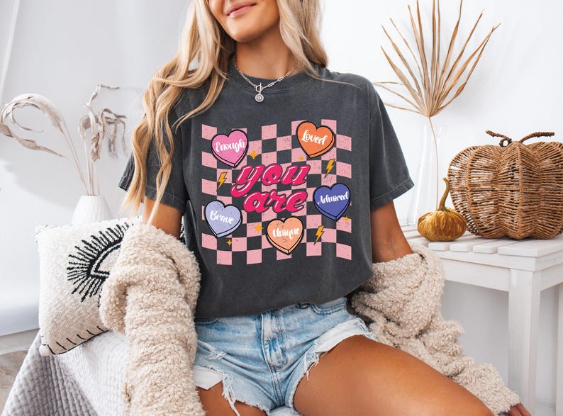 Puede incluir: Una camiseta gris oscuro recortada con un fondo a cuadros rosa y blanco. Las palabras "you are" est&aacute;n en rosa y rodeadas de cinco corazones con diferentes colores y palabras: "enough", "loved", "brave", "unique" y "adored".