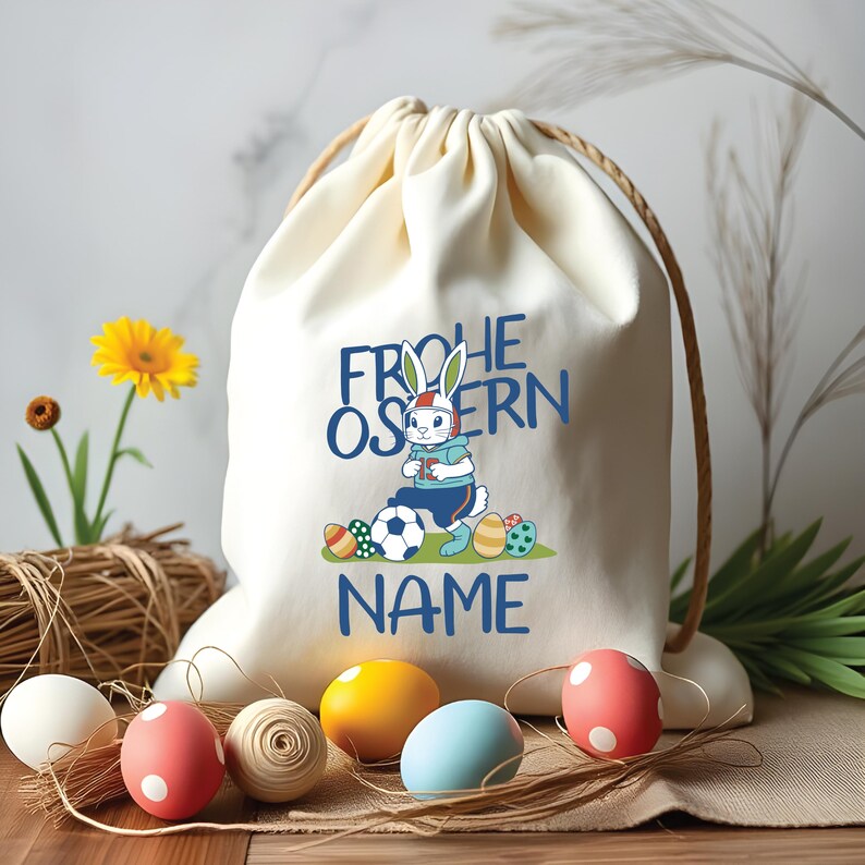 May include: A white canvas drawstring bag with a blue and white cartoon bunny wearing a helmet and holding a soccer ball. The bag has the text "Frohe Ostern" and "NAME" printed on it. The bag is surrounded by colorful Easter eggs.