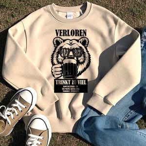 May include: A beige sweatshirt featuring a black graphic of a bear wearing sunglasses and holding a beer mug. The text "VERLOREN" is above the bear, and "TRINKT ZU VIEL" is below. Additional text is present. The sweatshirt is paired with jeans and sneakers.