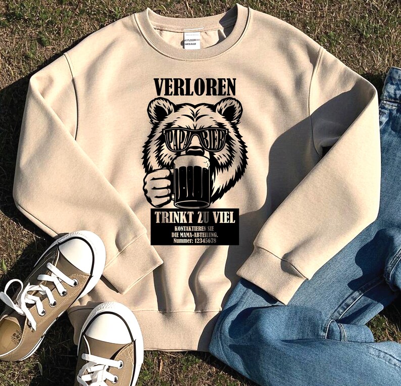 May include: A beige sweatshirt featuring a black graphic of a bear wearing sunglasses and holding a beer mug. The text "VERLOREN" is above the bear, and "TRINKT ZU VIEL" is below. Additional text is present. The sweatshirt is paired with jeans and sneakers.