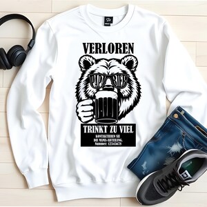 May include: White sweatshirt featuring a black and white bear graphic wearing sunglasses and holding a beer mug. The text on the sweatshirt reads "VERLOREN PAPA BIER TRINKT ZU VIEL".