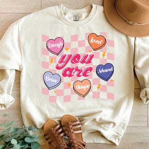 Puede incluir: Una sudadera de color crema con un fondo a cuadros rosa y blanco. Las palabras "you are" est&aacute;n en una fuente retro rosa y blanca. Cinco corazones rosas, morados y naranjas con palabras como "valiente", "&uacute;nico", "amado", "suficiente" y "admirado" est&aacute;n en el fondo a cuadros.