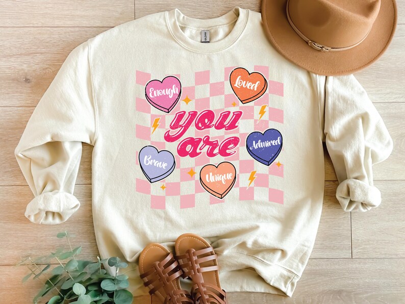 Puede incluir: Una sudadera de color crema con un fondo a cuadros rosa y blanco. Las palabras "you are" est&aacute;n en una fuente retro rosa y blanca. Cinco corazones rosas, morados y naranjas con palabras como "valiente", "&uacute;nico", "amado", "suficiente" y "admirado" est&aacute;n en el fondo a cuadros.