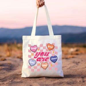 Puede incluir: Bolsa de tela blanca con un patr&oacute;n de cuadros rosa y blanco. La bolsa tiene cinco corazones de diferentes colores con palabras en ellos: "enough", "loved", "brave", "unique" y "adored". El texto "you are" est&aacute; escrito en una fuente retro rosa y blanca.