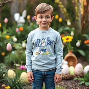 May include: A light blue long-sleeved sweatshirt with a graphic of a bunny rabbit playing soccer and the text "Frohe Ostern" in German.
