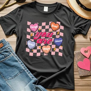 Puede incluir: Camiseta negra con un patr&oacute;n de cuadros rosa y negro y corazones con palabras como "amado", "suficiente", "valiente", "&uacute;nico" y "admirado". El texto "t&uacute; eres" est&aacute; en letras rosas grandes.