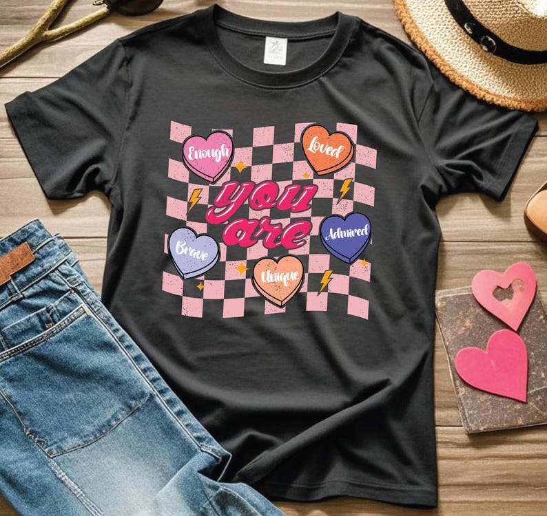 Puede incluir: Camiseta negra con un patr&oacute;n de cuadros rosa y negro y corazones con palabras como "amado", "suficiente", "valiente", "&uacute;nico" y "admirado". El texto "t&uacute; eres" est&aacute; en letras rosas grandes.