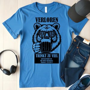 May include: A light blue t-shirt featuring a black bear graphic wearing sunglasses and holding a beer mug. The text on the shirt reads "VERLOREN PAPA BIER TRINKT ZU VIEL". A black baseball cap, blue jeans, and headphones are also visible.