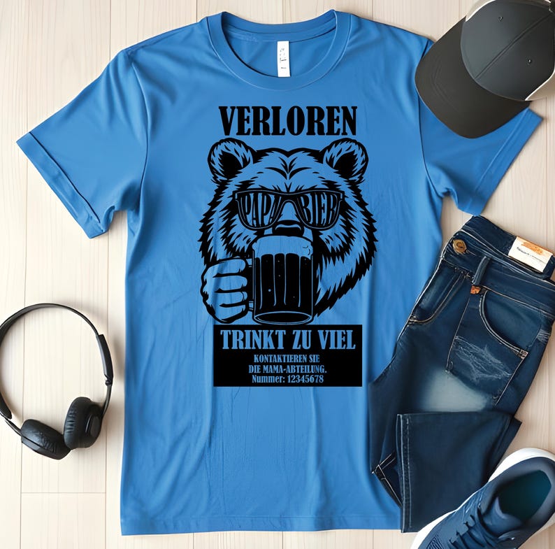 May include: A light blue t-shirt featuring a black bear graphic wearing sunglasses and holding a beer mug. The text on the shirt reads "VERLOREN PAPA BIER TRINKT ZU VIEL". A black baseball cap, blue jeans, and headphones are also visible.