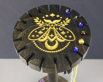 May include: A wooden jewelry stand with a black finish and a gold moth design. The stand has multiple slots for earrings and a post for necklaces.
