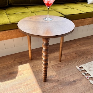 May include: A round wooden side table with a unique, twisted, wooden leg. The table has a dark brown wood finish and is sitting on a wood floor. A glass of red liquid with ice is sitting on the table.