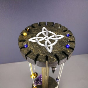 May include: A wooden stand with a Celtic knot design and slots for holding pendulums. The stand is dark brown with a silver-colored knot design. There are several pendulums hanging from the stand, some with blue and gold accents.