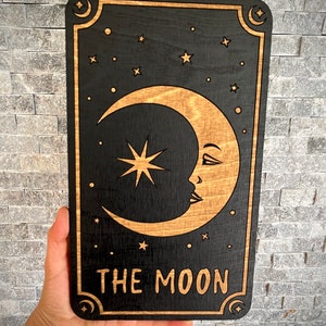 K&ouml;nnte beinhalten: Schwarze Tarotkarte aus Holz mit einem Halbmond mit Gesicht, einem Stern und anderen Sternen. Die Worte "THE MOON" sind unten eingraviert. Die Karte hat einen dekorativen Rand mit Monden und Sternen.