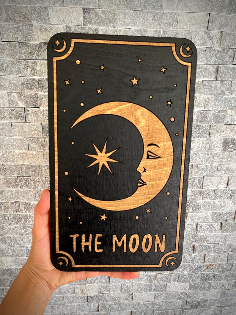 K&ouml;nnte beinhalten: Schwarze Tarotkarte aus Holz mit einem Halbmond mit Gesicht, einem Stern und anderen Sternen. Die Worte "THE MOON" sind unten eingraviert. Die Karte hat einen dekorativen Rand mit Monden und Sternen.