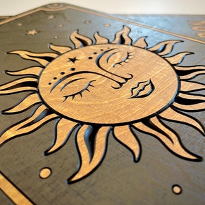 K&ouml;nnte beinhalten: H&ouml;lzerne Wandkunst mit einem Sonnen- und Monddesign. Die Sonne hat ein Gesicht mit geschlossenen Augen und einem Stern auf der Stirn. Der Hintergrund ist dunkelgrau mit Sternen. Sonne und Mond sind hellbraun.