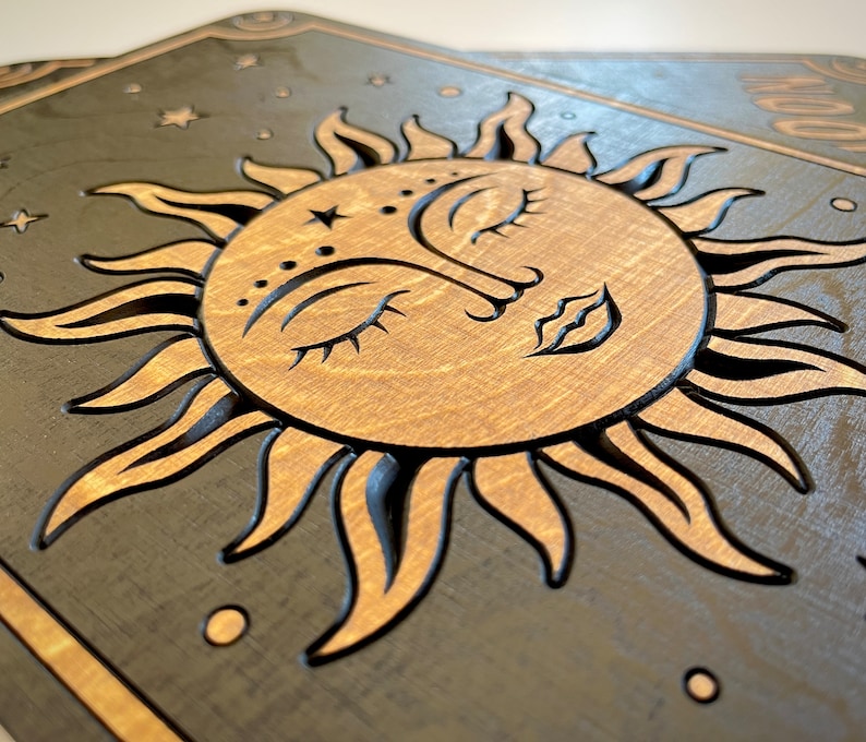 K&ouml;nnte beinhalten: H&ouml;lzerne Wandkunst mit einem Sonnen- und Monddesign. Die Sonne hat ein Gesicht mit geschlossenen Augen und einem Stern auf der Stirn. Der Hintergrund ist dunkelgrau mit Sternen. Sonne und Mond sind hellbraun.
