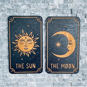 K&ouml;nnte beinhalten: Zwei Tarotkarten-Tafeln aus Holz, die Sonne und Mond darstellen. Die Sonnenkarte zeigt ein detailliertes Sonnendesign mit dem Text "THE SUN". Die Mondkarte zeigt einen Mond mit Gesicht und dem Text "THE MOON". Beide Karten sind schwarz mit goldenen Akzenten.