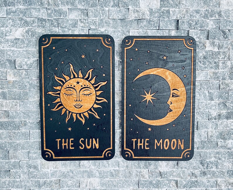 K&ouml;nnte beinhalten: Zwei Tarotkarten-Tafeln aus Holz, die Sonne und Mond darstellen. Die Sonnenkarte zeigt ein detailliertes Sonnendesign mit dem Text "THE SUN". Die Mondkarte zeigt einen Mond mit Gesicht und dem Text "THE MOON". Beide Karten sind schwarz mit goldenen Akzenten.