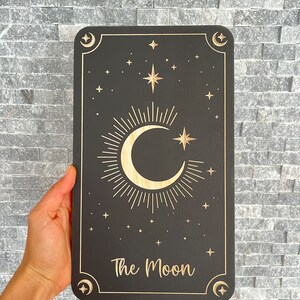 Może przedstawiać: Czarny drewniany znak z wzorem p&oacute;łksiężyca i gwiazd. Znak zawiera napis "The Moon" złotymi literami.