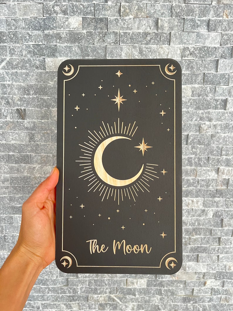 Może przedstawiać: Czarny drewniany znak z wzorem p&oacute;łksiężyca i gwiazd. Znak zawiera napis "The Moon" złotymi literami.