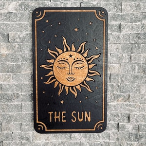 K&ouml;nnte beinhalten: Ein Holzschild mit einem Sonnenmotiv, das ein l&auml;chelndes Gesicht mit geschlossenen Augen zeigt. Die Sonne ist hellbraun auf schwarzem Hintergrund. Die Worte "THE SUN" stehen unten. Das Schild ist rechteckig mit abgerundeten Ecken.