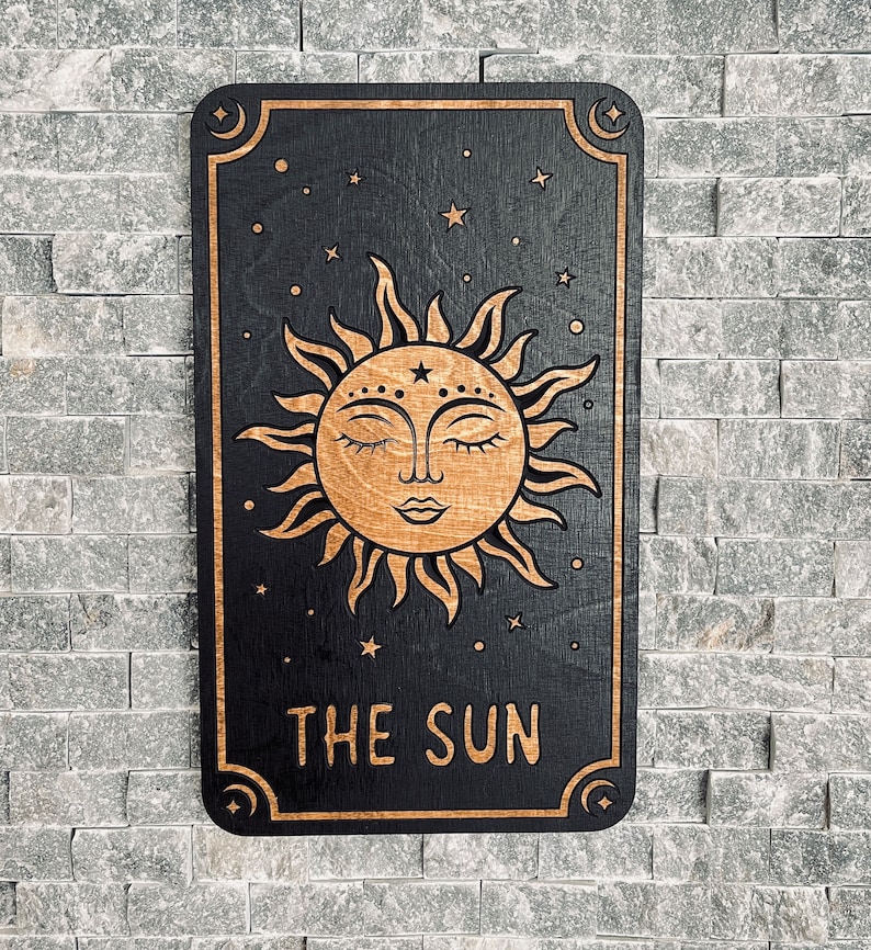 K&ouml;nnte beinhalten: Ein Holzschild mit einem Sonnenmotiv, das ein l&auml;chelndes Gesicht mit geschlossenen Augen zeigt. Die Sonne ist hellbraun auf schwarzem Hintergrund. Die Worte "THE SUN" stehen unten. Das Schild ist rechteckig mit abgerundeten Ecken.