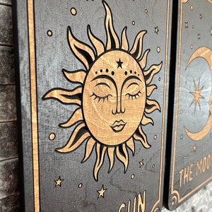 K&ouml;nnte beinhalten: Wandkunst aus Holz mit einem Sonnen- und Monddesign. Die Sonne hat ein l&auml;chelndes Gesicht mit geschlossenen Augen und ist von Strahlen umgeben. Der Mond hat eine Sichelform mit einem Stern darin. Der Hintergrund ist dunkelgrau mit goldenen Akzenten.