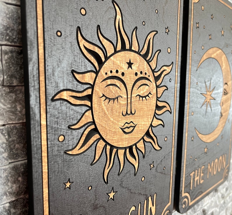 K&ouml;nnte beinhalten: Wandkunst aus Holz mit einem Sonnen- und Monddesign. Die Sonne hat ein l&auml;chelndes Gesicht mit geschlossenen Augen und ist von Strahlen umgeben. Der Mond hat eine Sichelform mit einem Stern darin. Der Hintergrund ist dunkelgrau mit goldenen Akzenten.