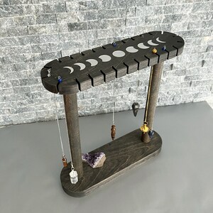 May include: A wooden stand with a moon phase design, used to display pendulums and crystals. The stand has a dark wood finish and is oval shaped. It has 12 slots for hanging pendulums and a flat surface for displaying crystals.