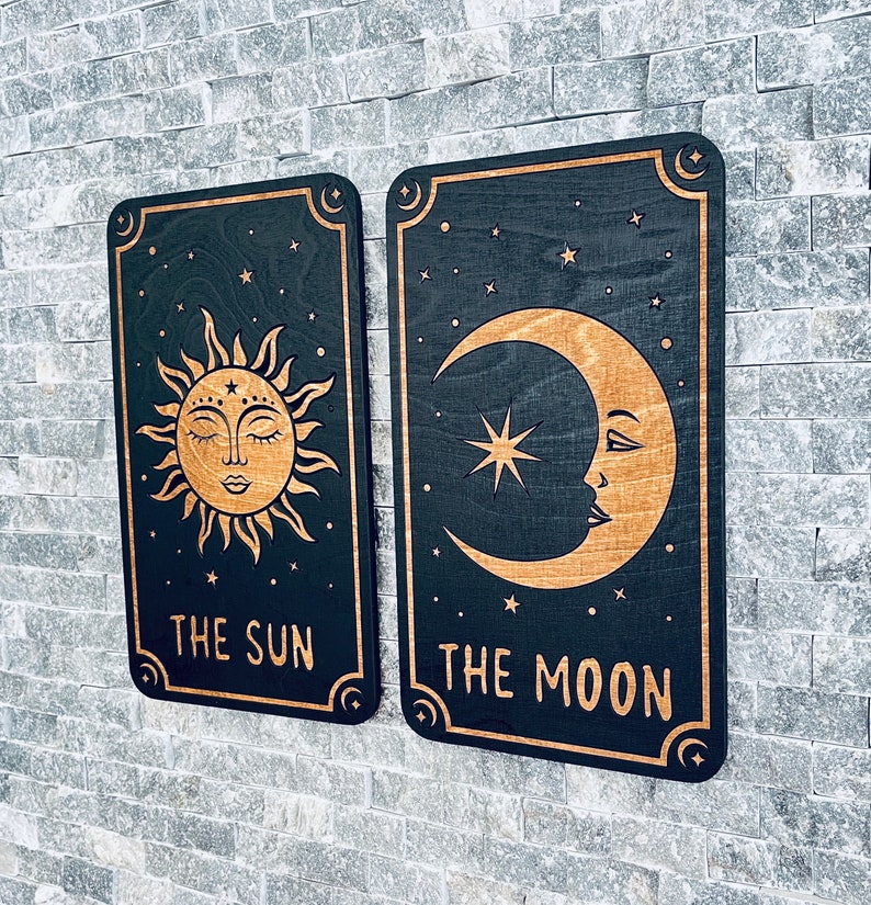 K&ouml;nnte beinhalten: Zwei Wandbeh&auml;nge aus Holz im Tarotkarten-Design. Einer zeigt eine Sonne mit geschlossenen Augen, der andere einen Mond und einen Stern. Beide sind schwarz mit goldfarbenen Designs und dem Text: "THE SUN" und "THE MOON".